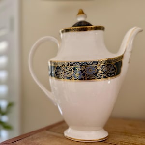 May include: A white porcelain teapot with a curved handle and spout. The teapot has a dark teal band with gold floral detailing and gold trim. The lid is topped with a gold finial. The teapot sits on a wooden surface.