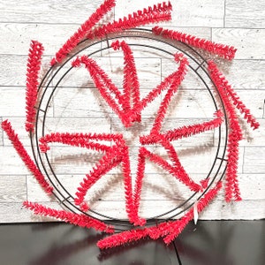 May include: A red and black wreath with a wire frame and red tinsel. The tinsel is arranged in a starburst pattern.