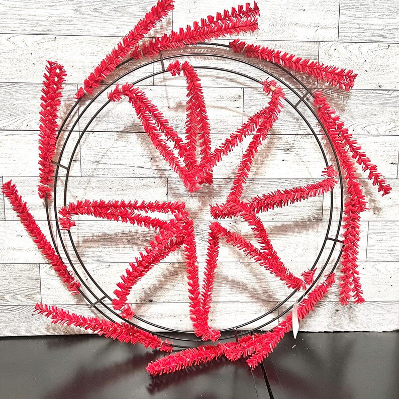 May include: A red and black wreath with a wire frame and red tinsel. The tinsel is arranged in a starburst pattern.