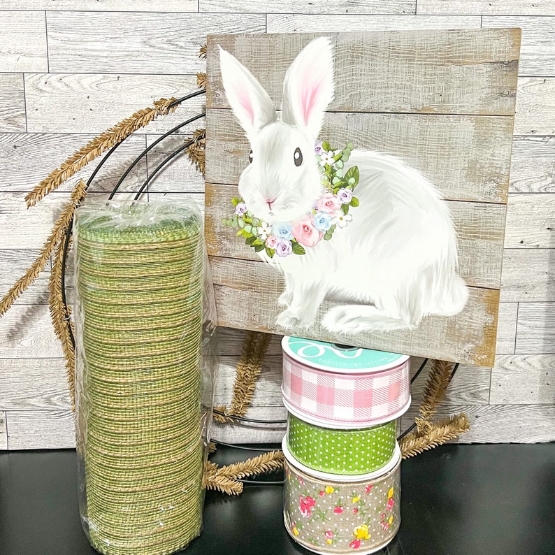 May include: A green burlap ribbon roll, a stack of three decorative ribbons, and a wooden sign with a white bunny wearing a floral wreath. The bunny is on a woodgrain background.