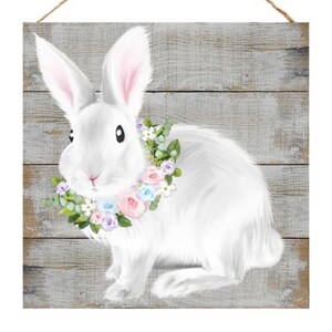 May include: A white rabbit with a pink, blue, and white flower crown sits on a gray wooden background.