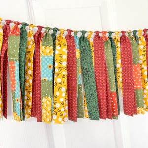 May include: A colorful fabric garland with alternating strips of patterned fabric. The garland features red, green, yellow, and blue fabrics with floral and polka dot designs. The garland is hanging against a white background.