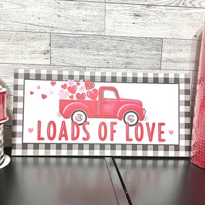 May include: A red truck with a white bed is filled with pink and red hearts. The truck is on a black and white chequered background with the text "Loads of Love" in red.