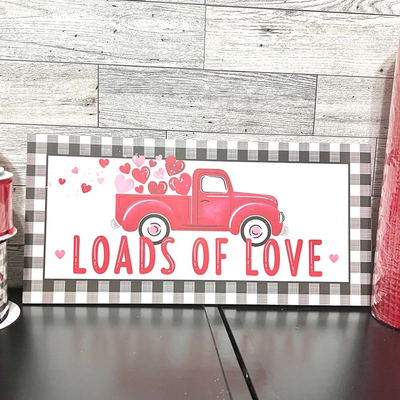 May include: A red truck with a white bed is filled with pink and red hearts. The truck is on a black and white chequered background with the text "Loads of Love" in red.