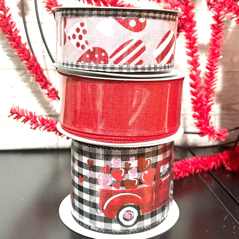 May include: Three rolls of ribbon with Valentine's Day themes. The top roll is white with red, pink, and black polka dots and stripes. The middle roll is red. The bottom roll is black and white gingham with red hearts and a red truck with white hearts.
