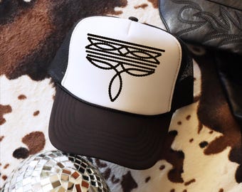May include: A trucker hat with a white front panel and a black mesh back. The hat features a black graphic design on the front, with a brown brim and a black button on top. The hat is a stylish accessory.