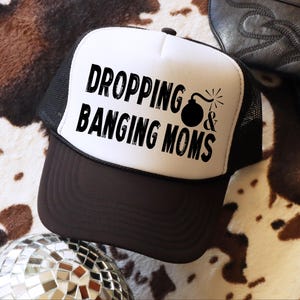 May include: A black and white trucker hat with the text "DROPPING & BANGING MOMS" in black. The hat has a brown brim and a black mesh back. A disco ball and a cowboy boot are in the background.