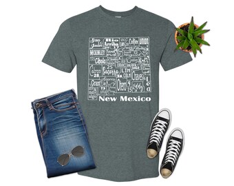 May include: A dark green t-shirt with a white graphic of the state of New Mexico. The graphic is made up of the names of the counties in the state, arranged in a map-like shape. The text "New Mexico" is printed below the graphic.