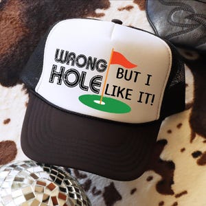May include: A black and white trucker hat with the text "WRONG HOLE BUT I LIKE IT!". The hat features a white front panel with a golf-themed graphic and a brown brim. The hat has a black mesh back.