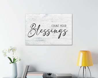 May include: White wood sign with black text that reads "Count Your Blessings".