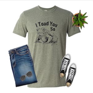 May include: A grey t-shirt with the phrase "I Toad You So" and a black toad illustration. The image also features blue denim jeans, black trainers, sunglasses, and a small potted plant.