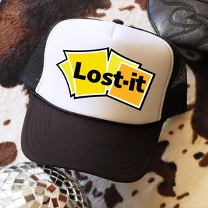 May include: A trucker hat with a white front panel and a black mesh back. The front features the text "Lost-it" in black letters on yellow and orange sticky notes. The hat has a brown peak and a black button on top.