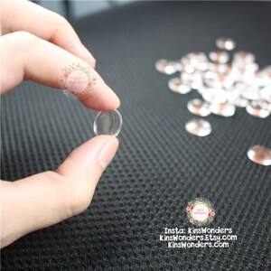 May include: A close-up of a clear glass cabochon, approximately 1 inch in diameter, held in a hand. The cabochon is round and smooth, with a slight dome shape. The cabochon is surrounded by a scattering of similar cabochons on a black textured surface.