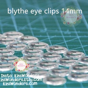 May include: A close-up of a group of clear plastic eye clips for Blythe dolls. The clips are 14mm in diameter and are arranged in rows on a blue cutting mat. The text "blythe eye clips 14mm" is visible at the top of the image.