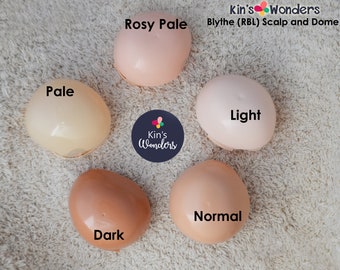 May include: Five plastic doll head sculpts in different skin tones: Rosy Pale, Pale, Light, Normal, and Dark. The sculpts are arranged in a circle on a white fluffy surface. The text "Kin's Wonders Blythe (RBL) Scalp and Dome" is at the top of the image.