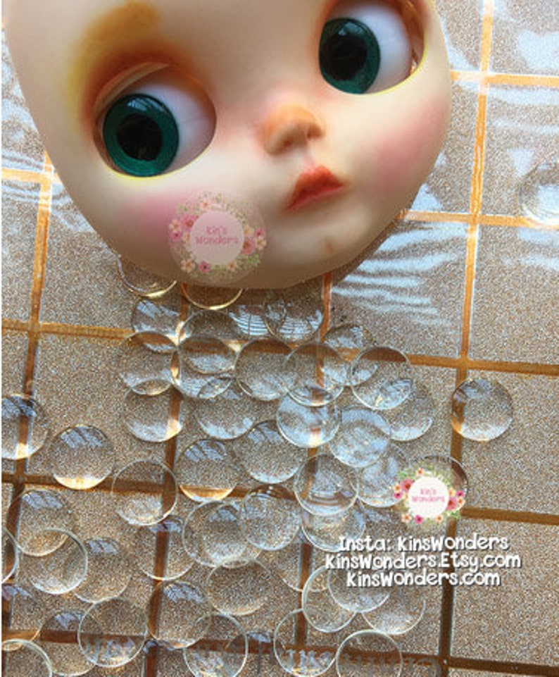 May include: A close-up of a doll's face with green eyes and a pink blush. The doll is surrounded by a large quantity of clear glass or plastic round embellishments.