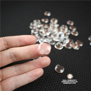 May include: A close-up of a pile of clear round glass cabochons, with one cabochon being held by a finger. The cabochons are about 1/2 inch in diameter.
