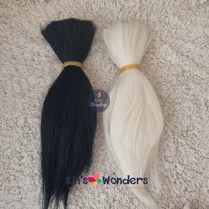 Puede incluir: Dos mechones de pelo de muñeca, uno negro y otro blanco, atados con gomas elásticas. El pelo es largo y liso. Los mechones están sobre una superficie blanca y peluda.