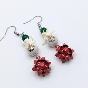 May include: A pair of dangle earrings featuring red bow charms, white and gold beads, and green beads. The earrings have silver-colored hooks. The earrings are on a white background.