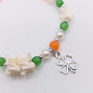 May include: A beaded bracelet featuring white, green, and orange beads, along with small, off-white decorative elements. A silver charm in the shape of a four-leaf clover dangles from the bracelet. The bracelet is on a white background.
