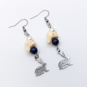 May include: A pair of dangle earrings featuring a silver bunny charm with a floral design. Each earring has a cream-colored stone, a dark blue bead, and a white pearl. The earrings hang from silver hooks.