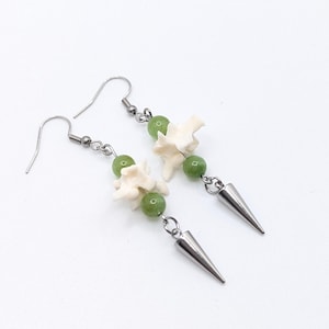 May include: A pair of dangle earrings featuring white bone-shaped beads, green beads, and silver-colored cone-shaped charms. The earrings have silver-colored ear wires and connecting chains. The jewelry is displayed against a white background.
