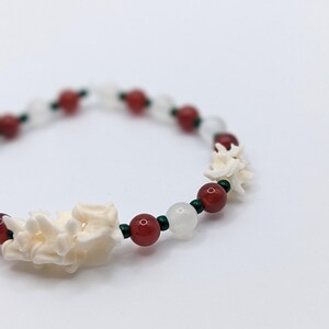 May include: A beaded bracelet featuring alternating white floral-shaped beads, red round beads, and white round beads. Small green beads separate the other beads. The bracelet is on a white background.