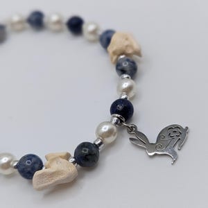 May include: A beaded bracelet featuring white faux pearls, dark blue beads, and small, off-white bone-shaped beads. A silver-colored charm in the shape of a rabbit with a flower detail hangs from the bracelet. The bracelet is on a white background.