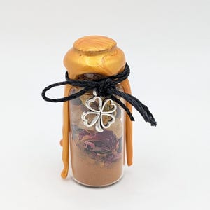 May include: A small clear glass bottle filled with brown powder, dried rose petals, and light-colored stones. The bottle is sealed with a gold-colored wax stopper and tied with a black string featuring a silver-toned four-leaf clover charm.
