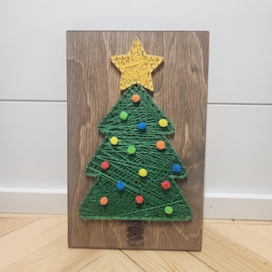 May include: A handmade Christmas tree string art piece. The tree is crafted with green string on a wooden board, adorned with colorful pom-poms. A yellow star tops the tree. The wooden board has a brown finish.