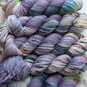 May include: Close-up of several skeins of hand-dyed yarn in shades of lavender, teal, and cream. The yarn is arranged in a loose, textured pattern, with visible color variations. The yarn is suitable for knitting and crochet projects.