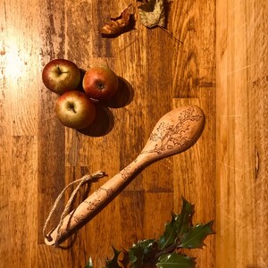 May include: A wooden spoon with a carved holly design, three red apples, and a sprig of holly with green leaves on a wooden surface.