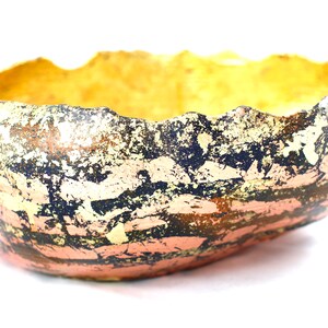 Könnte beinhalten: Eine handgefertigte Keramikschale mit einer strukturierten, unebenen Oberfläche. Die Schale ist mit goldener und schwarzer Farbe verziert, was ein einzigartiges und abstraktes Design ergibt.