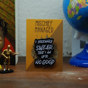Könnte beinhalten: Eine schwarz-weiße Emaille-Anstecknadel mit dem Text "Mischief Managed" und "I solemnly swear that I am up to no good". Die Anstecknadel befindet sich auf einer weißen Karte mit schwarzem Rand.