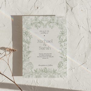 Op de afbeelding: Een trouwuitnodiging met een lichtgroene bloemenrand. De tekst luidt "join us for the wedding of Michael & Sarah" met datum, tijd en locatie eronder. De uitnodiging staat op een witte, getextureerde achtergrond.