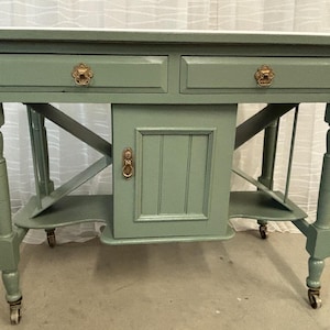 May include: Sage green antique table with two drawers, a cabinet door, and decorative brass hardware. The table has turned legs and a lower shelf, and it sits on small caster wheels. The table's dimensions are approximately 36 inches wide, 18 inches deep, and 30 inches high.