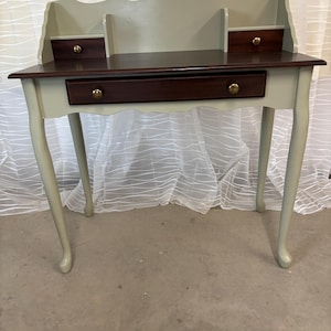May include: A small, light green desk with a dark brown top. The desk has a single drawer and two small shelves on the back. The desk has four legs with a curved design.