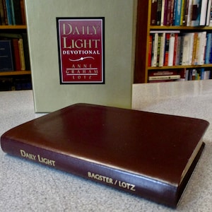 May include: A green and brown leather-bound book titled "Daily Light Devotional" by Anne Graham Lotz. The book is closed and sitting on a table.
