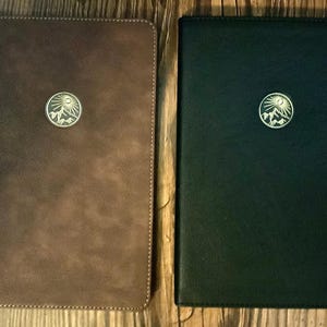 May include: Two leather-bound journals, one brown and one black, are displayed side-by-side. Each journal features a circular emblem with a mountain and sun design. The journals are rectangular with rounded corners and stitched edges.