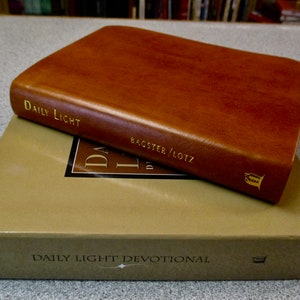 May include: A brown leather-bound book titled "Daily Light" by  "Bagster/Lotz" with gold lettering. The book is sitting on a box with the text "Daily Light Devotional" printed on the front.