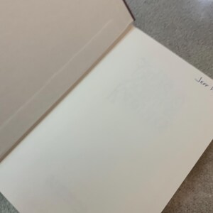 Puede incluir: Libro abierto con p&aacute;ginas blanquecinas y una cubierta burdeos. La p&aacute;gina derecha tiene un texto tenue y una inscripci&oacute;n manuscrita en tinta azul que dice "Jerr F". El libro est&aacute; abierto sobre una superficie gris.