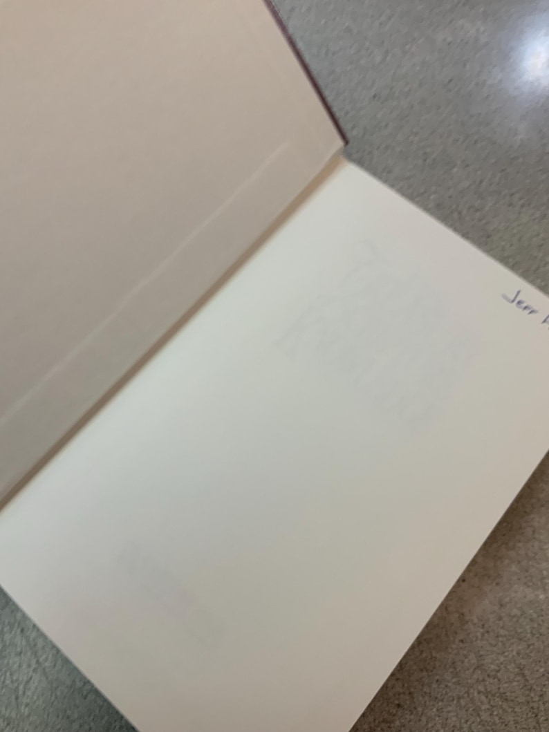 Puede incluir: Libro abierto con p&aacute;ginas blanquecinas y una cubierta burdeos. La p&aacute;gina derecha tiene un texto tenue y una inscripci&oacute;n manuscrita en tinta azul que dice "Jerr F". El libro est&aacute; abierto sobre una superficie gris.