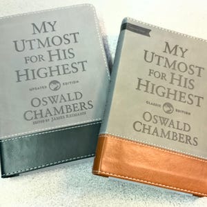 May include: Two books titled My Utmost for His Highest by Oswald Chambers. The left book has a gray cover with a dark charcoal bottom panel. The right book has a gray cover with a tan bottom panel. Both feature white stitching and embossed text.