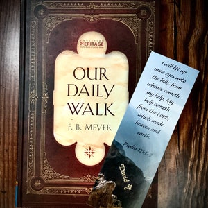 May include: A brown leather-bound book titled "Our Daily Walk" by F.B. Meyer. The book has a gold-colored design on the cover and a bookmark with a mountain scene and a quote from Psalm 121:1-2.