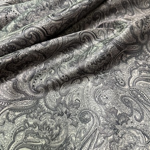 May include: Close-up of a fabric with a gray and white paisley pattern. The fabric is draped, showing the intricate design of swirling teardrop shapes and floral motifs. The color palette is monochromatic, with varying shades of gray.
