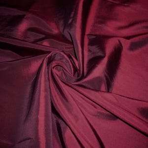 Może przedstawiać: Zbliżenie na głęboką bordową tkaninę o lśniącym połysku. Materiał jest zebrany i zakręcony, tworząc fałdy i cienie, które podkreślają teksturę i wariacje kolorystyczne. Tkanina wydaje się gładka i płynna.