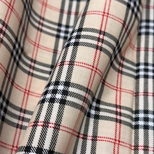 May include: Close-up of beige fabric with a classic plaid pattern. The design features black, white, and red lines creating a grid-like arrangement. The fabric is folded, showing the texture and drape of the material. Ideal for apparel and home decor.