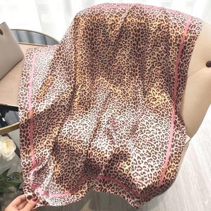 Può includere: Una sciarpa quadrata di seta con stampa leopardata nei toni del marrone e crema. La sciarpa ha un bordo rosa ed è drappeggiata su una sedia. Il tessuto è morbido e fluido.