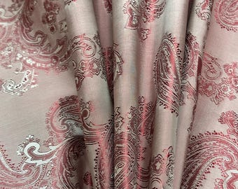 May include: Close-up of a fabric with a paisley pattern. The fabric is a light pink color with a repeating design in a darker rose hue. The fabric is draped, showing folds and texture.