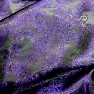 Pode incluir: Tecido roxo com padrão paisley e brilho acetinado. O tecido está dobrado e drapeado, mostrando a textura e o detalhe do padrão.
